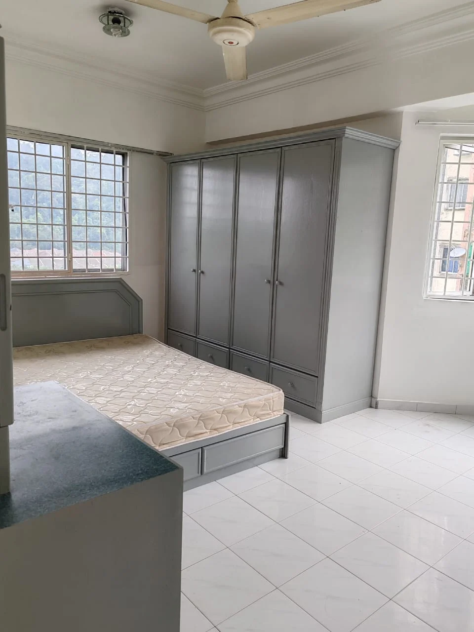 Bedroom features: queen-size bed with mattress, built-in grey wardrobe with sliding doors, white painted walls, white tiled flooring, ceiling fan, large window with grid pattern, natural light from window, corner of room with a small cabinet; style: modern; view: mountain.